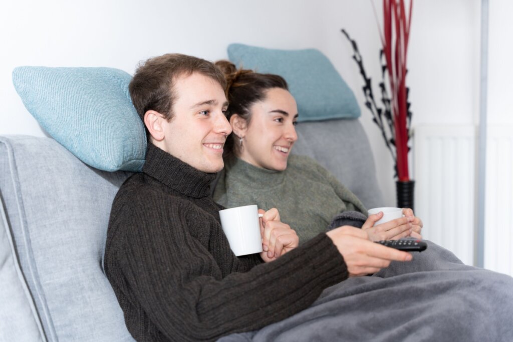 Casal sentado no sofá vendo TV, com cobertas e canecas nas mãos