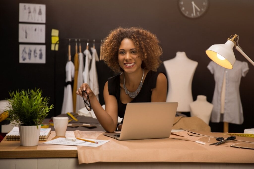 Mulher sentada em uma mesa com computador e elementos de costura ao fundo