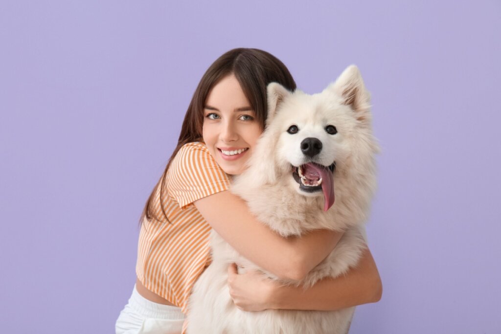 menina de camiseta laranja e abraçando cachorro samoieda branco em fundo roxo