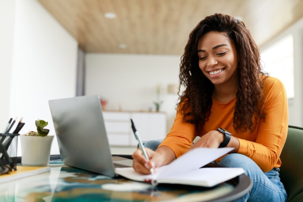 menina sorrindo estudando com notebook e caderno em cima de uma mesa