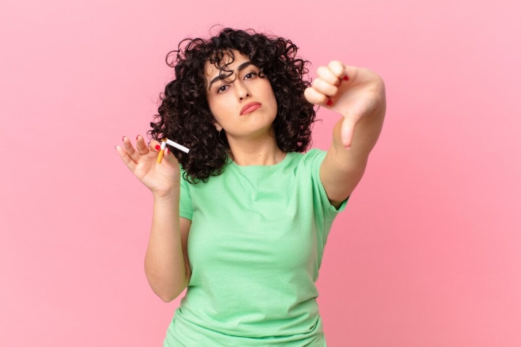 mulher segurando cigarro com uma mão e fazendo sinal de reprovação com a outra mão em fundo rosa