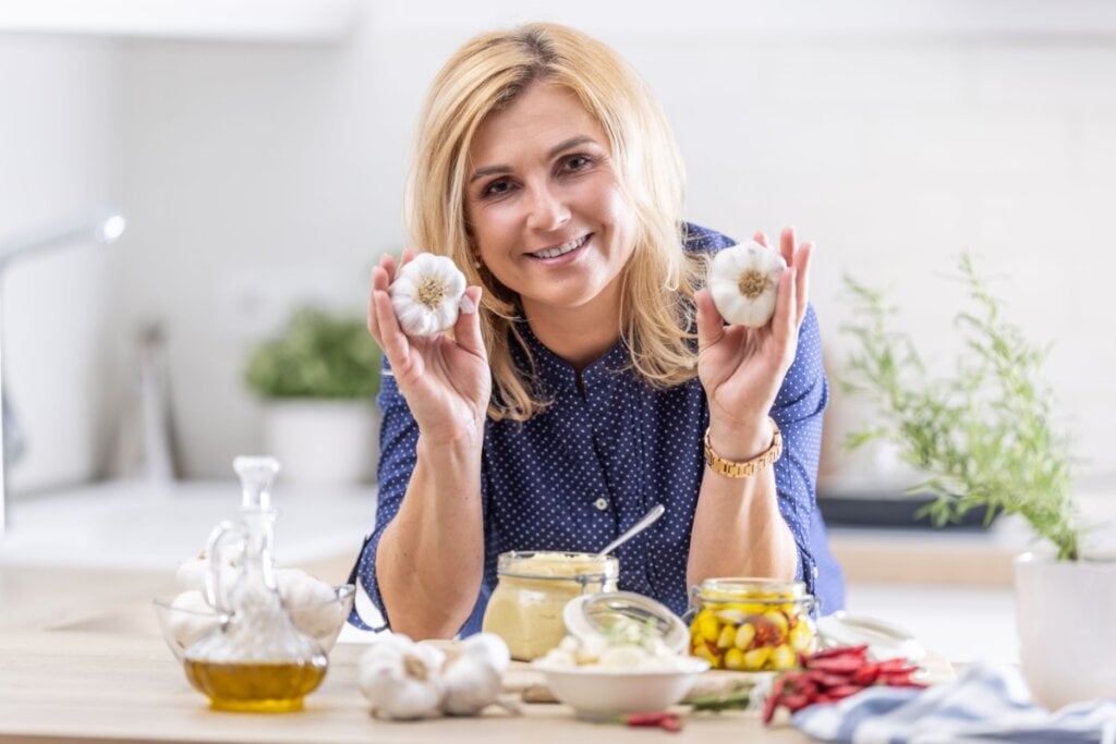 Mulher em cozinha preparando alho de diversas formas e segurando alho em cada mão