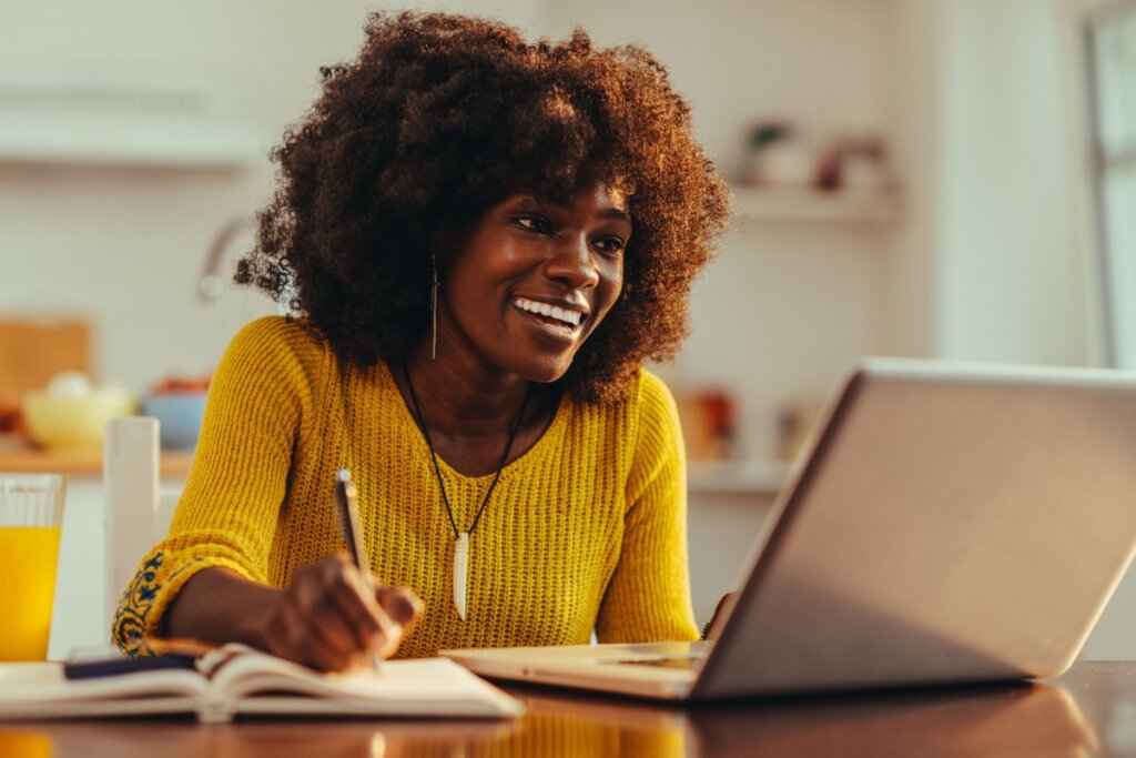 Mulher jovem escrevendo em caderno e sorrindo em frente a um notebook