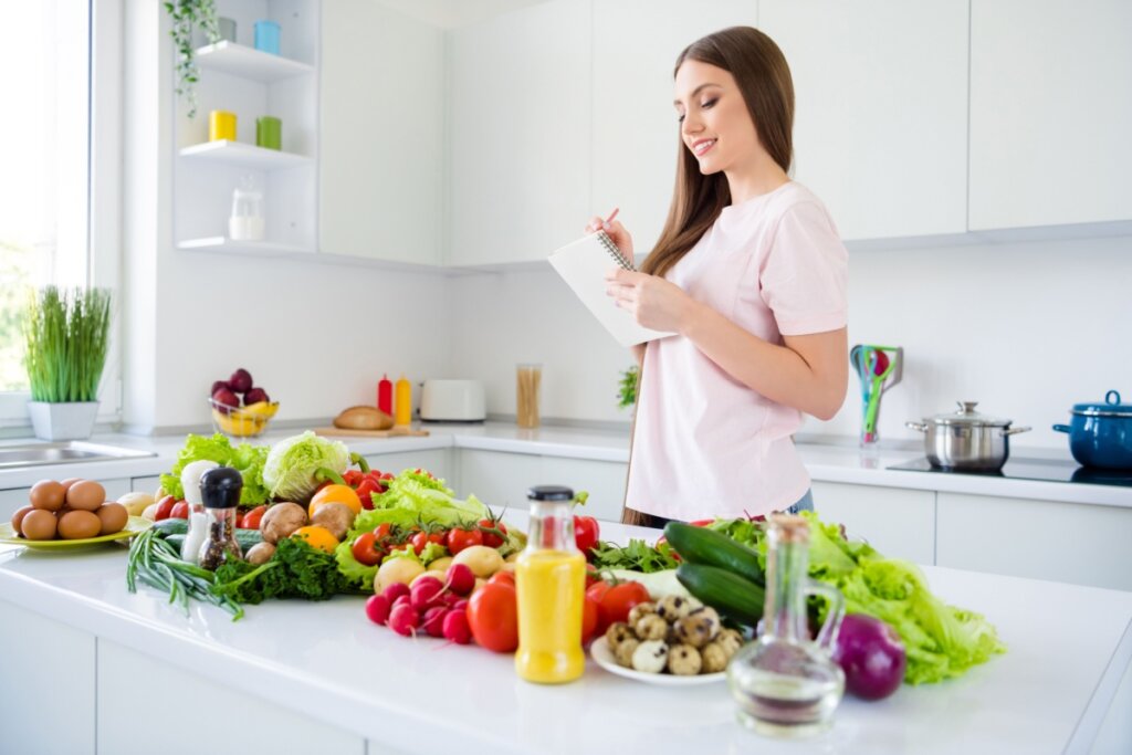 Mulher na cozinha fazendo uma lista e com diversos alimentos saudáveis espalhados na bancada