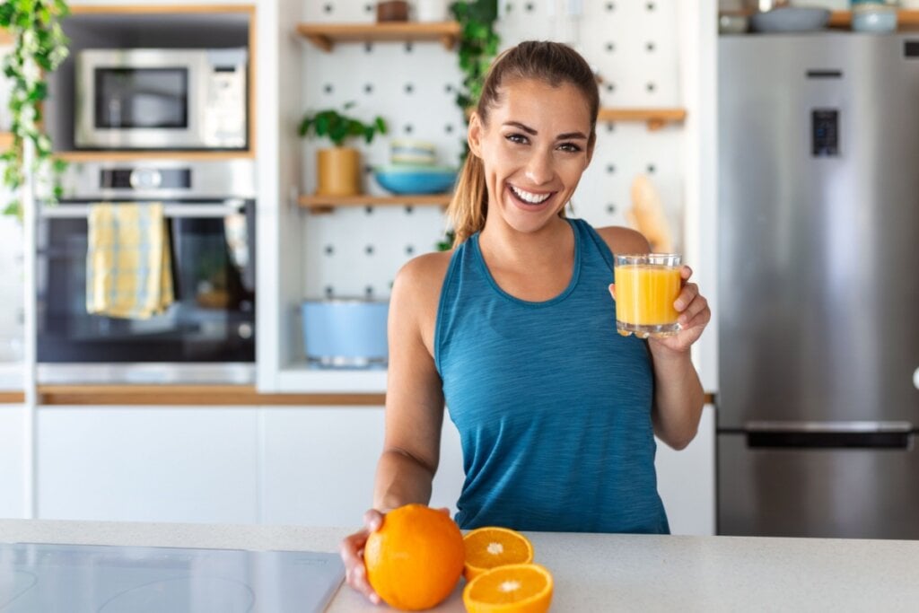 Mulher bebendo suco de laranja encostada em uma bancada na cozinha, com laranjas na bancada