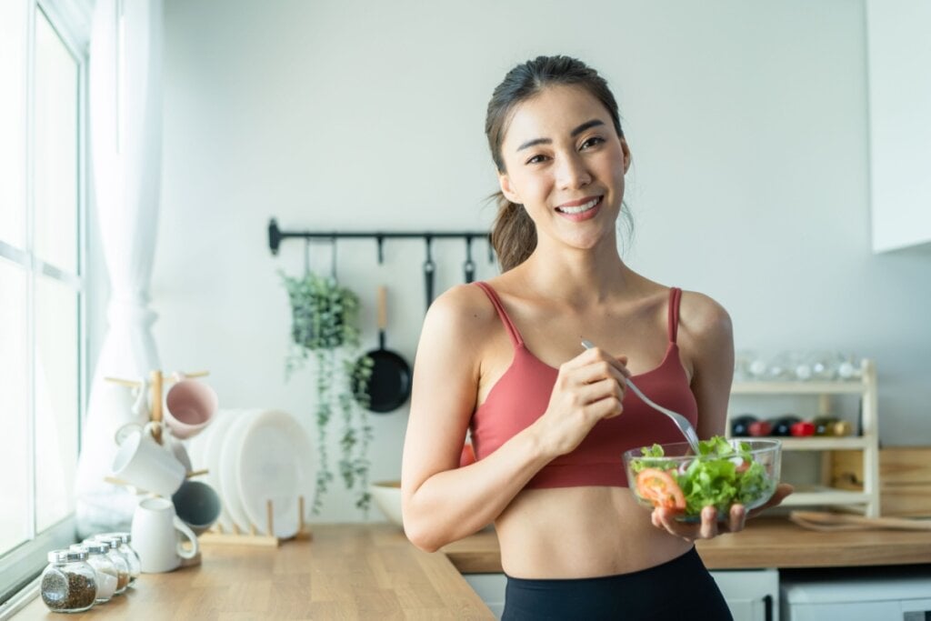 Mulher na cozinha segurando pote de salada e garfo na mão