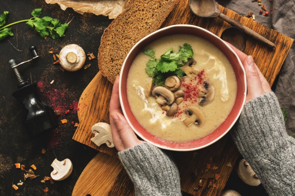 Tigela de sopa cremosa de cogumelos, decorada com cogumelos frescos e folhas de salsinha, sendo segurada por uma pessoa vestindo um suéter de lã cinza