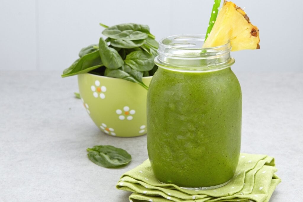 Suco verde em copo de vidro com canudo verde e fatia de abacaxi. Pote verde com espinafre ao fundo