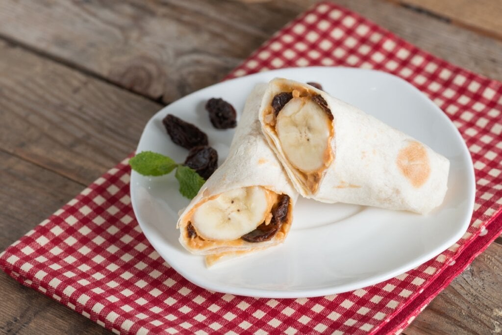 Tortilha de banana, pasta de amendoim e uvas-passas cortada em cima de prato branco