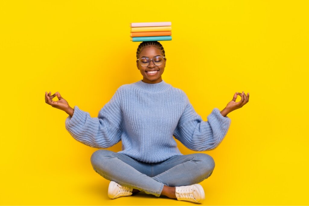 Mulher sentada com as pernas cruzadas em frente a um fundo amarelo meditando com livros na cabeça