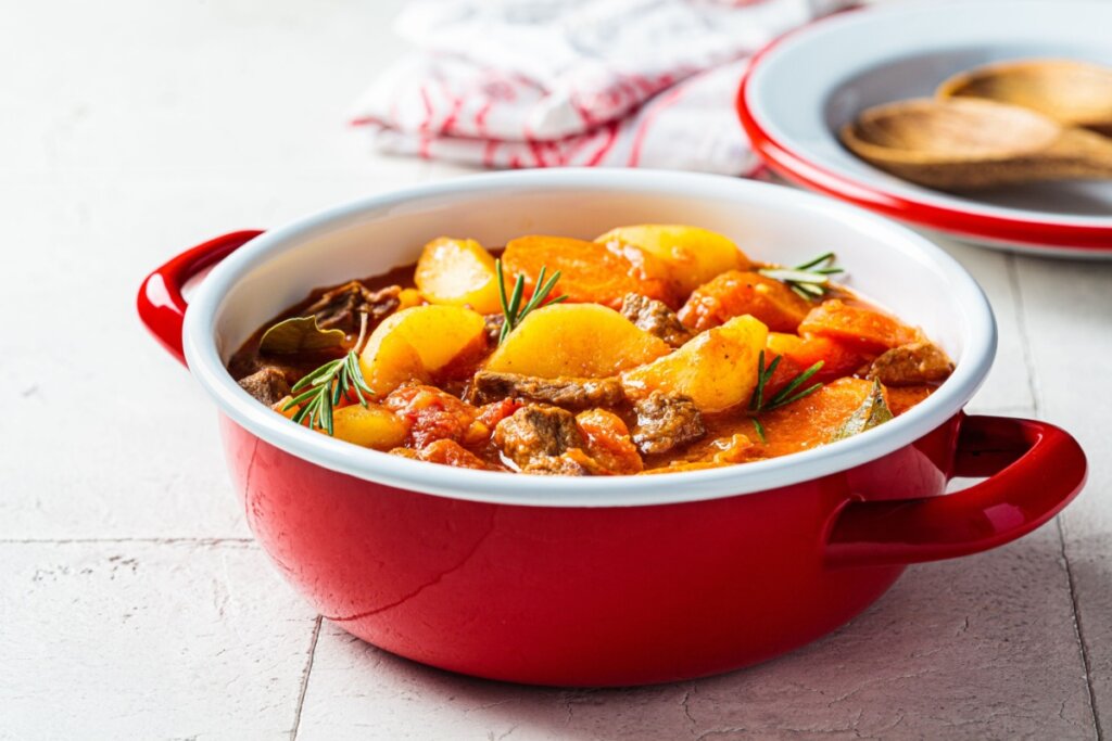 Carne de panela com legumes em panela vermelha em cima de mesa branca