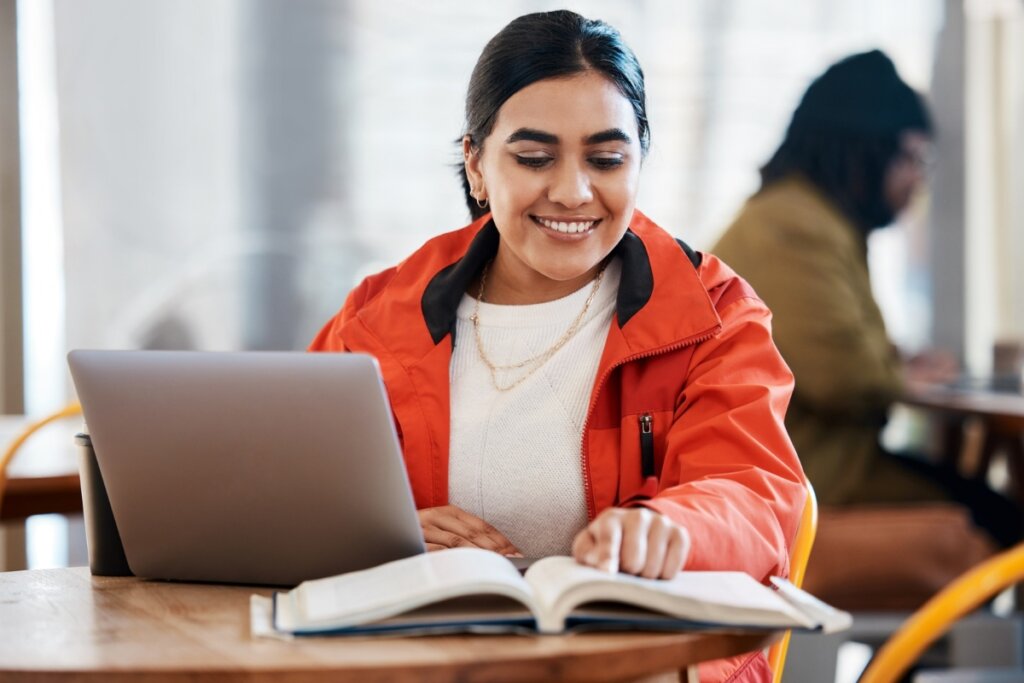 Mulher sentada em frente a um notebook e lendo um livro em cima de uma mesa