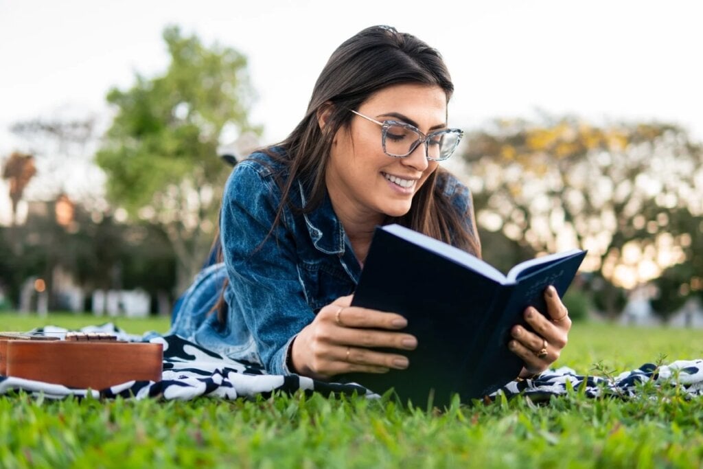 Mulher deitada na grama de uma jardim lendo um livro