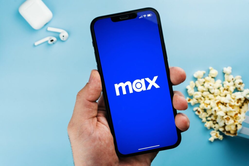 Logo do Max em um celular com uma pessoa segurando e um fone e balde de pipoca ao lado