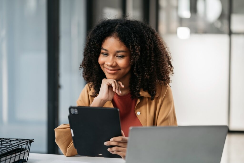 Mulher sorrindo, sentada em frente a um notebook mexendo em um tablet