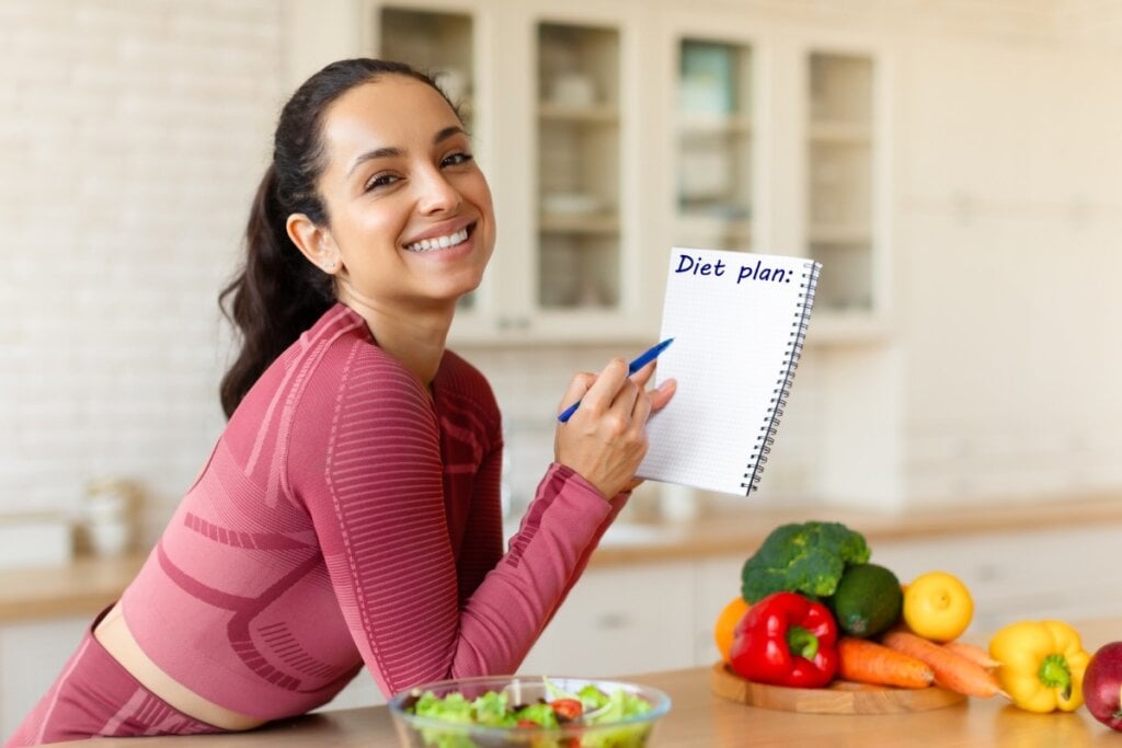 Mulher sorrindo apoiada em uma bancada na cozinha segurando um caderno 