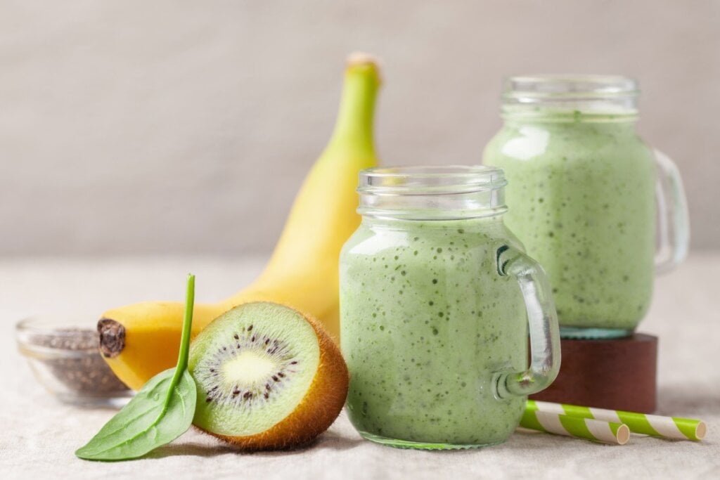 Shake de banana, kiwi, espinafre e chia em dois copos transparentes de vidro; banana, chia, folha verde, chia e canudos ao lado