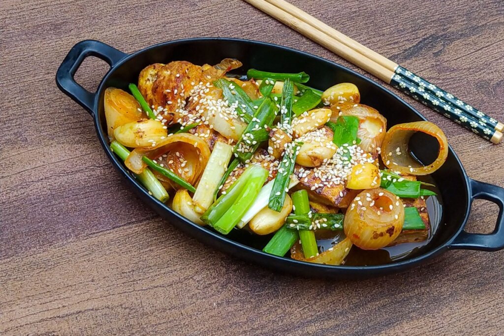 Tofu grelhado com alho e molho coreano, servido com cebolinha, sementes de gergelim e cebolas caramelizadas em uma travessa preta com hashis ao lado