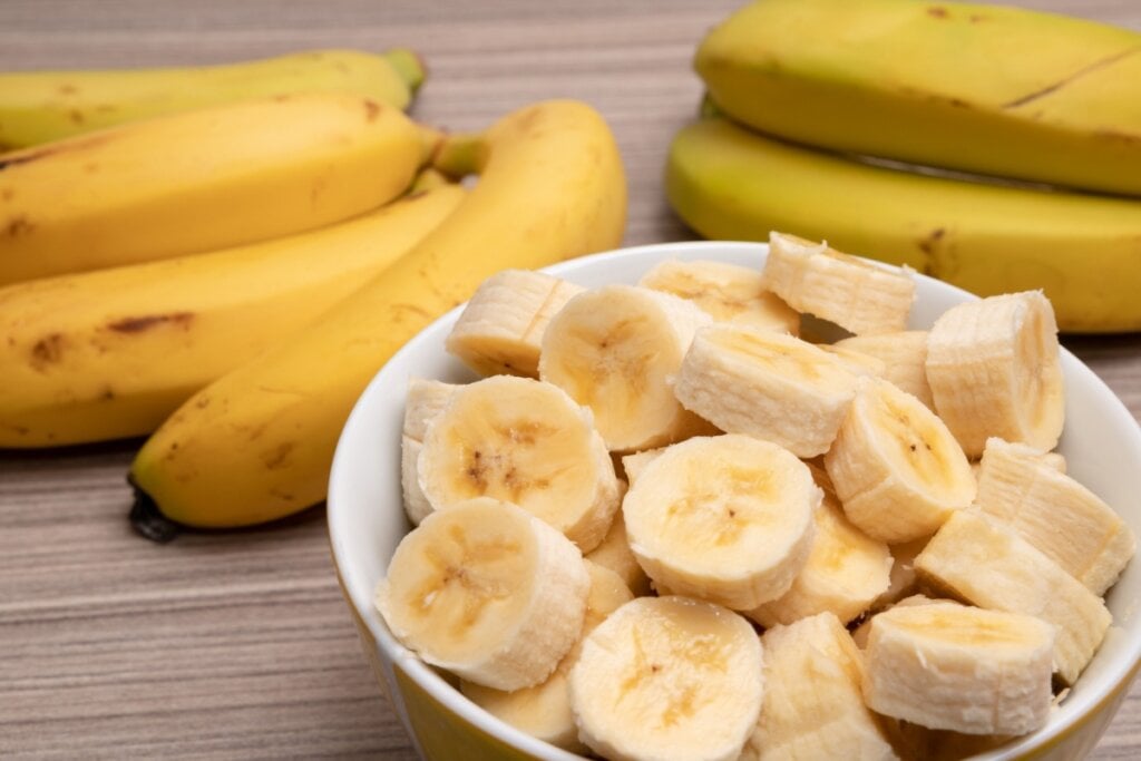 Banana cortada em rodelas em uma tigela branca com bananas ao redor em cima de mesa de madeira