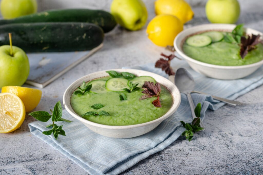 creme verde de pepino em recipiente branco e pano azul. pepino, limão, maçã-verde e hortelã em volta