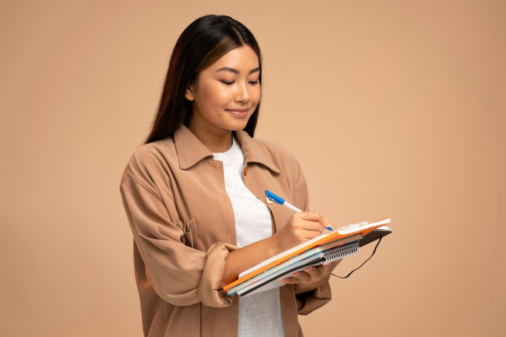 estudante de blusa marrom escrevendo em caderno que ela segura com uma das mãos