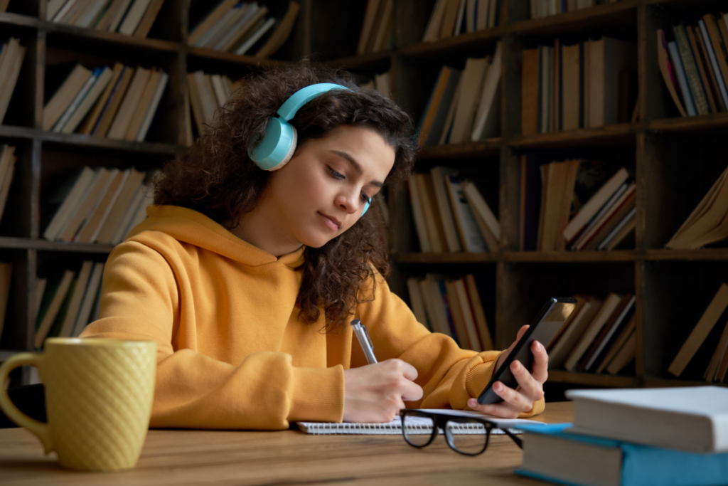 menina em ambiente com livro sentada escrevendo em caderno olhando o celular