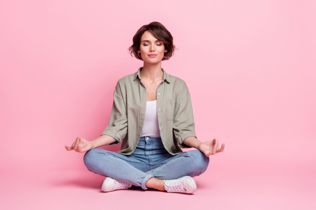 mulher sentada no chão com as pernas cruzadas, os olhos fechados e meditando