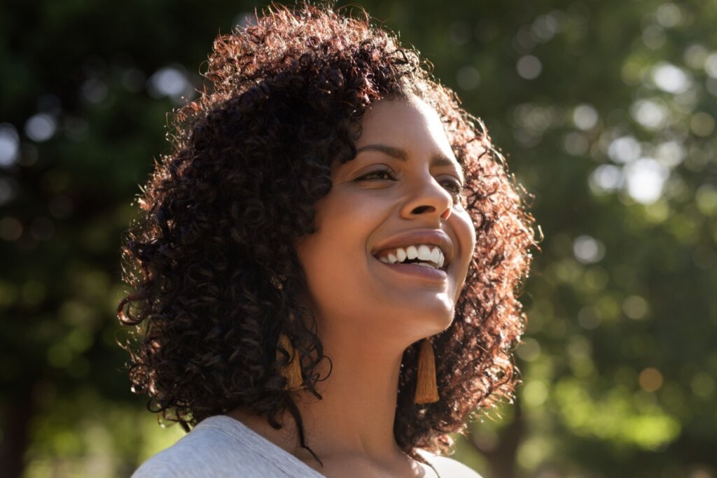 mulher de cabelo cacheado sorrindo e com árvores ao fundo