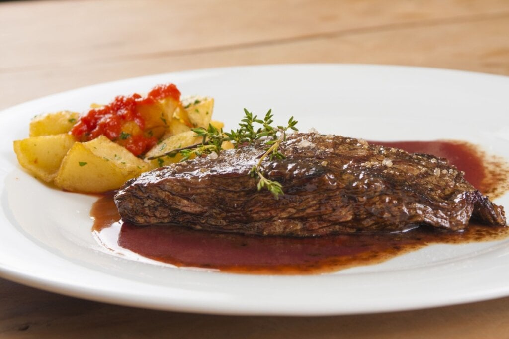 Picanha com molho de vinho tinto servido em prato branco com batatas ao lado