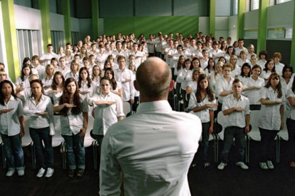 Homem em um palco e alunos em baixo o imitando no filme 