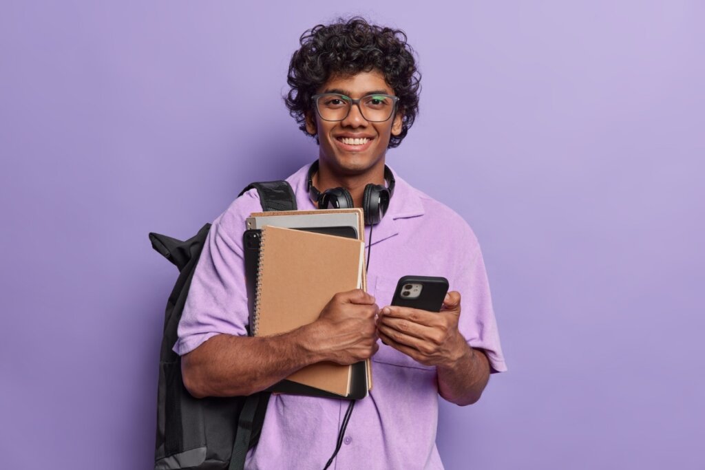 Menino usando óculos de grau e camisa roxa segurando cadernos em uma mão e um celular na outra