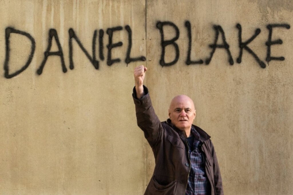 Homem em frente a uma parede com grafite com um dos braços levantados para o alto em cena do filme "Eu, Daniel Blake" 