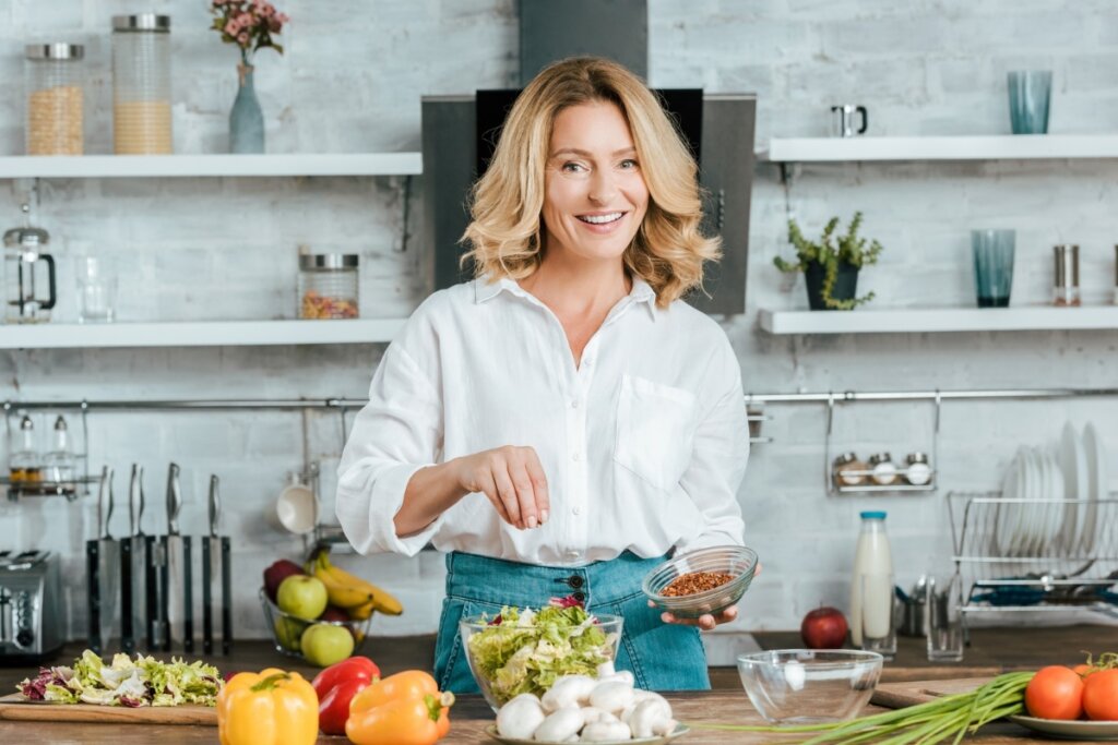 Mulher usando camisa branca em uma cozinha em frente a uma bancada com alimentos