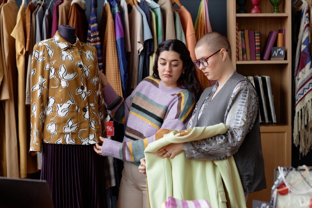 Mulher mexendo em uma roupa em um manequim ao lado de outra mulher segurando um caderno