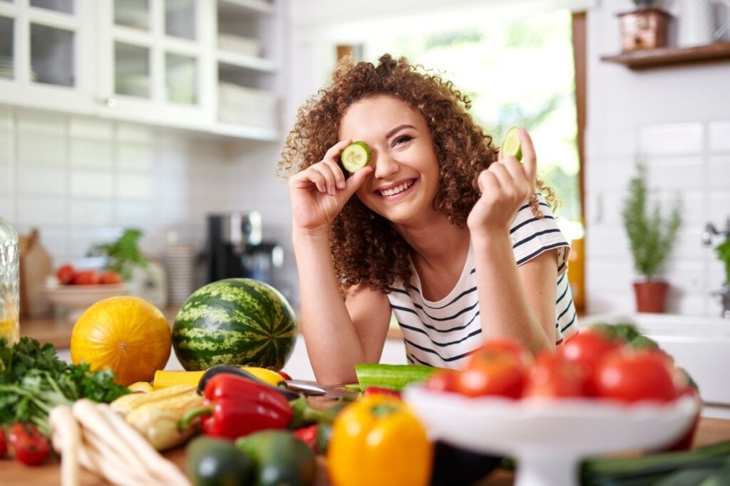 Mulher sorrindo em frente a uma bancada com alimentos e segurando uma rodela de pepino em frente ao olho