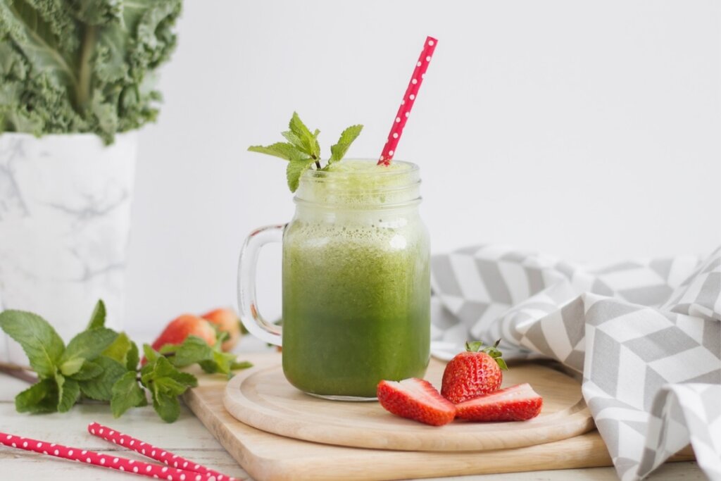 Caneca de vidro com suco verde decorado com um canudo vermelho em cima de uma tábua de madeira com pedaços de morango