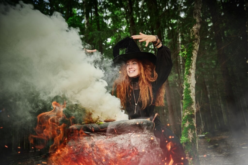 Mulher vestida como bruxa segurando uma vassoura e usando um chapéu pontiagudo, ao lado de um caldeirão soltando fumaça