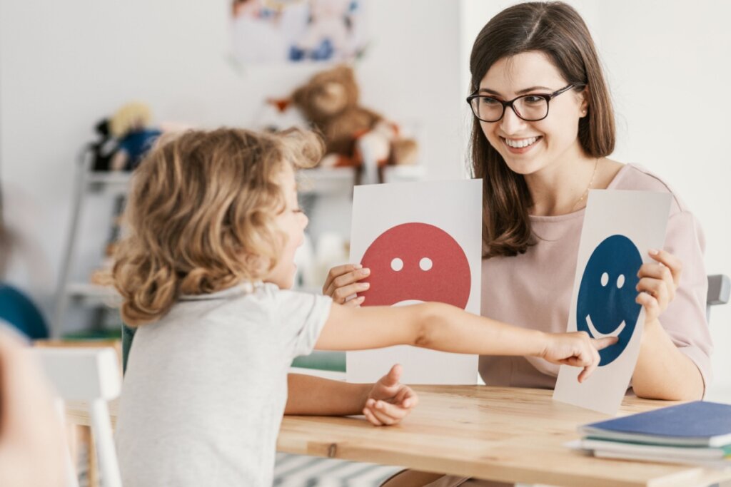 psicóloga segurando duas folha com emoji, uma feliz e outra triste. Criança apontando para a feliz