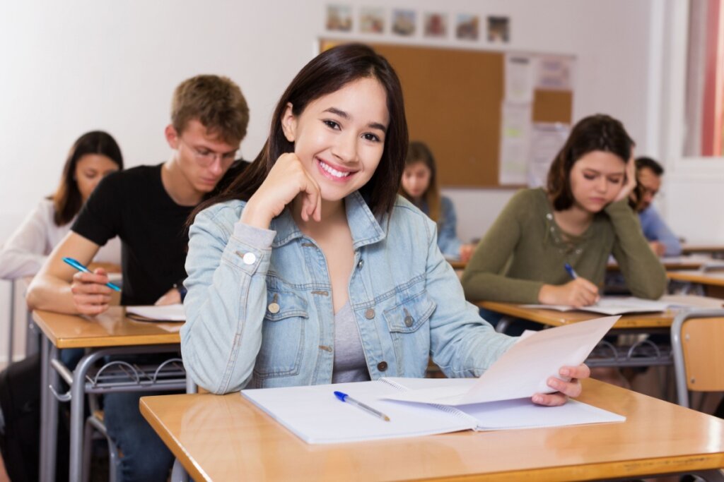 estudante em sala de aula, sentada, com prova na mesa e sorrindo