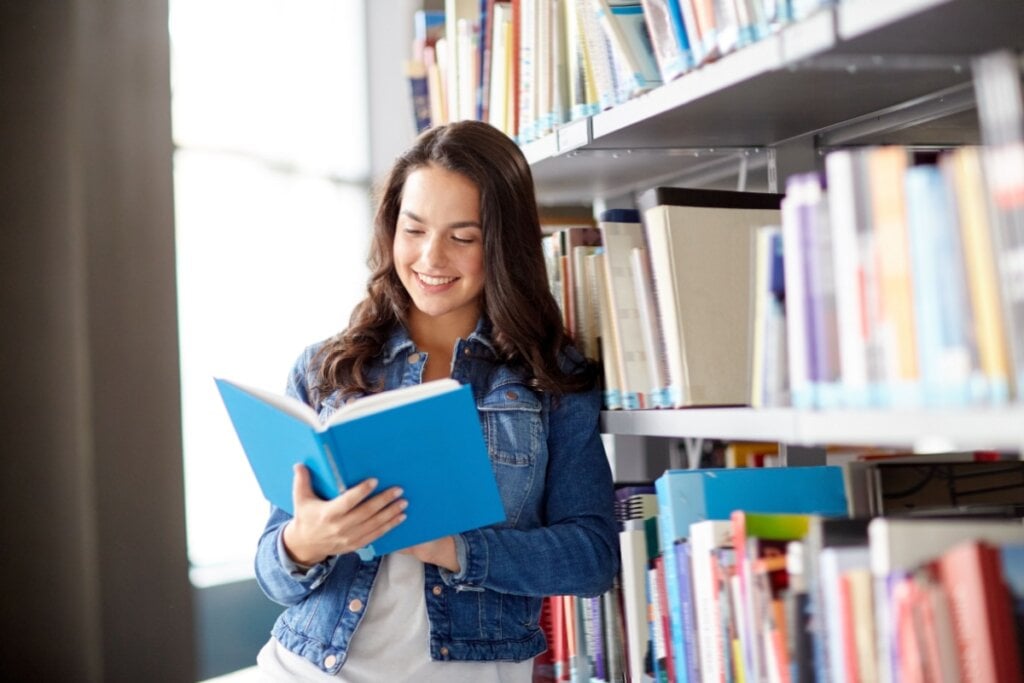 menina lendo livro de capa azul e apoiada em estante de biblioteca com livros