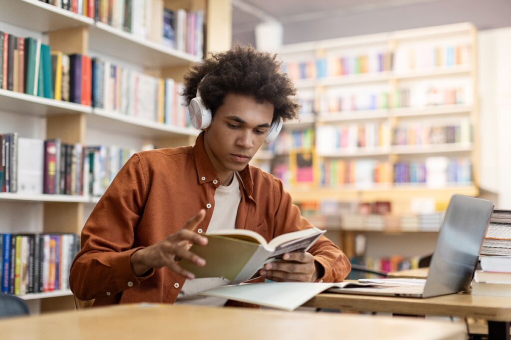 Estudante em biblioteca lendo livro com notebook ao lado e fone de ouvido