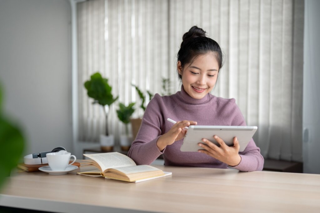 menina sentada e mexendo em tablet e livro aberto sobre mesa