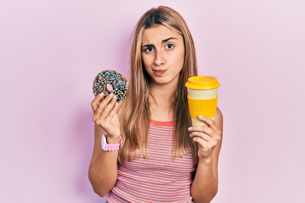 Jovem segurando um donnut e uma bebida, um em cada mão em um fundo rosa