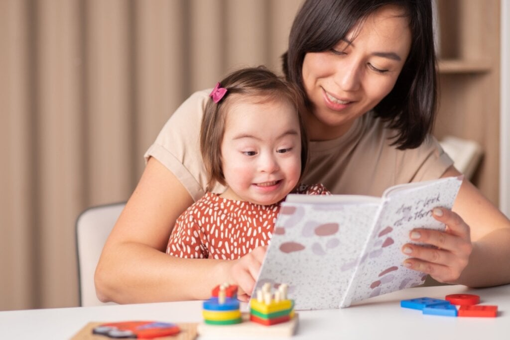 mulher com criança com Síndrome de Down no colo lendo e mostrando o livro pra ela