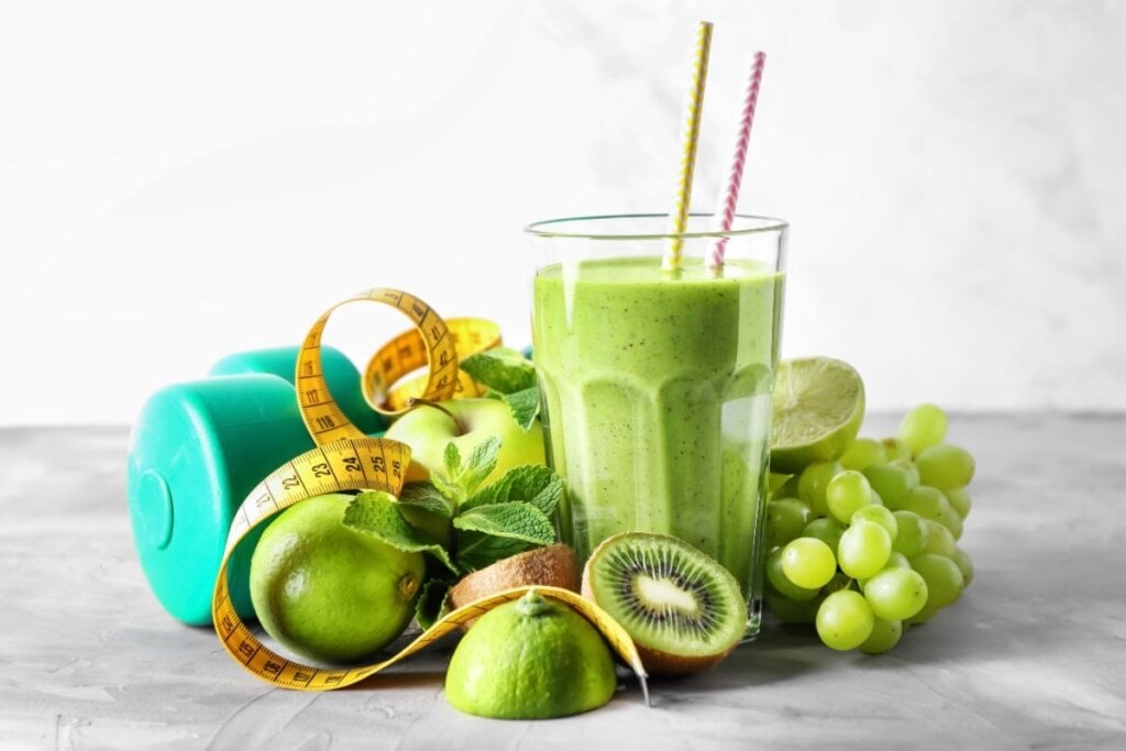 Suco verde em um copo de vidro cercado por frutas frescas, incluindo kiwi, uva, limão e maçã, acompanhado por um haltere e uma fita métrica