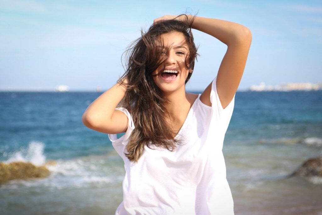 Mulher usando blusa branca em uma praia com as duas mãos no cabelo