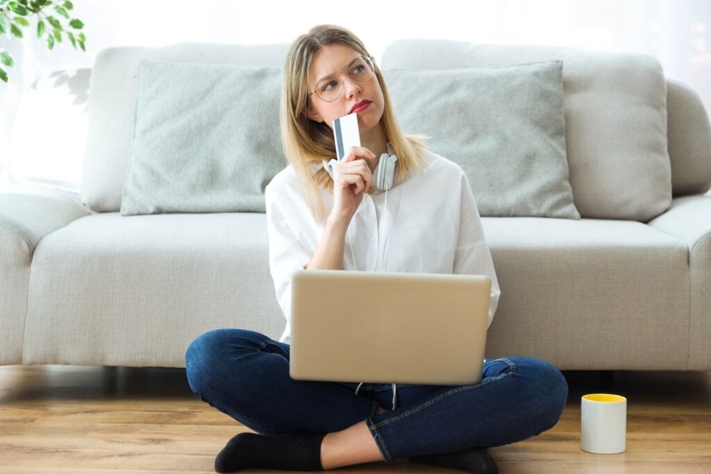 Mulher sentada com um notebook no colo, olhando para o lado e segurando um cartão próximo ao queixo como se estivesse pensando