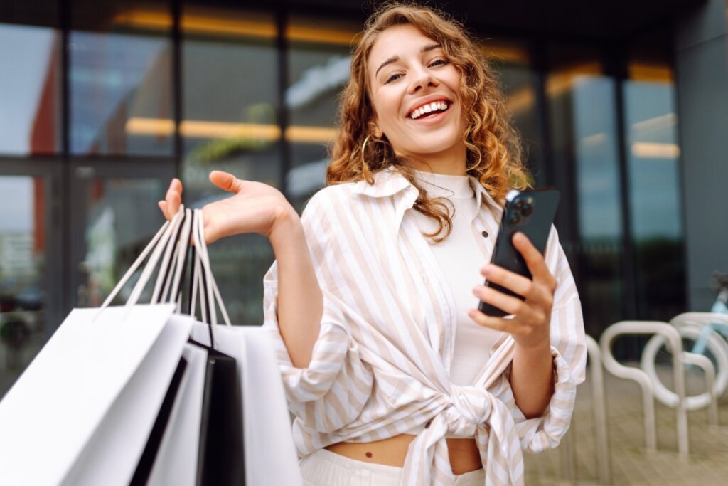 Mulher sorrindo segurando sacolas de compras em uma mão e o celular em outra