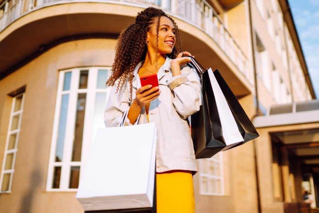 Mulher na rua segurando sacolas de compras em uma mão e um celular na outra