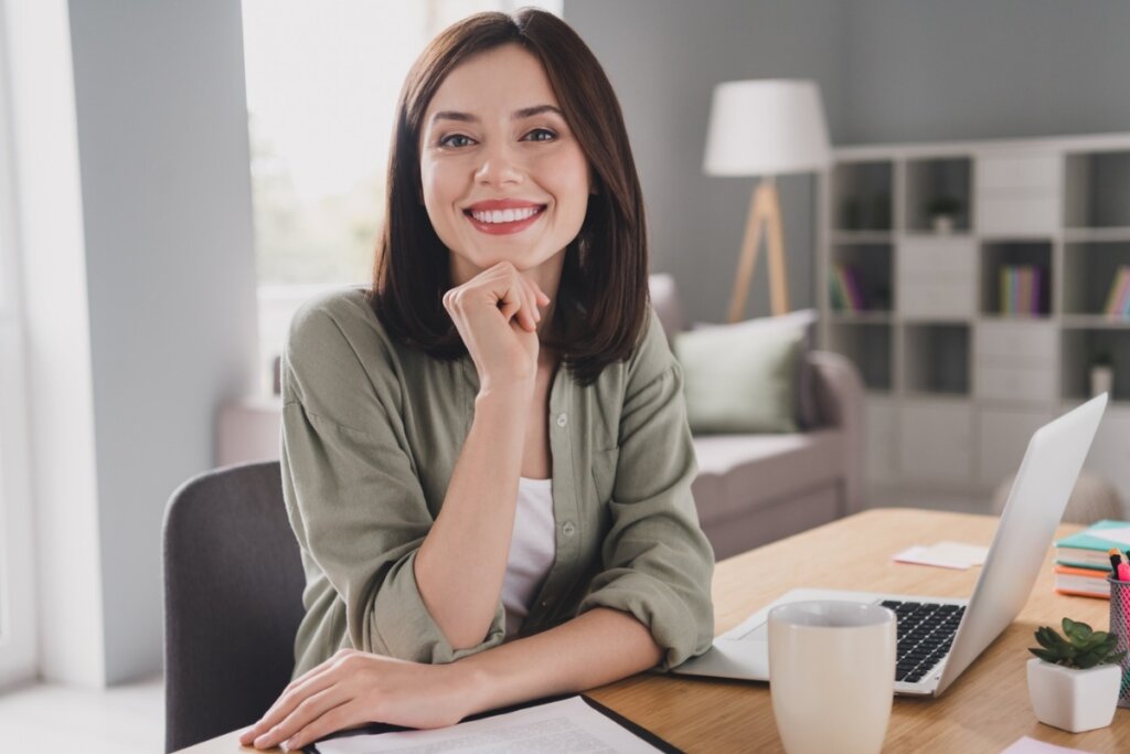 Mulher sentada com a mão no queixo e sorrindo em frente a uma mesa com um notebook e papéis